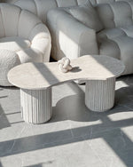 Table basse en travertin naturel aux pieds cylindriques striés, mise en scène dans un salon bohème chic avec canapé beige, tapis crème et plante verte.
